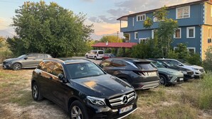 Un estacionamiento con varios automóviles, incluyendo un SUV Mercedes-Benz negro, y un edificio de dos pisos con techo rojo al fondo.