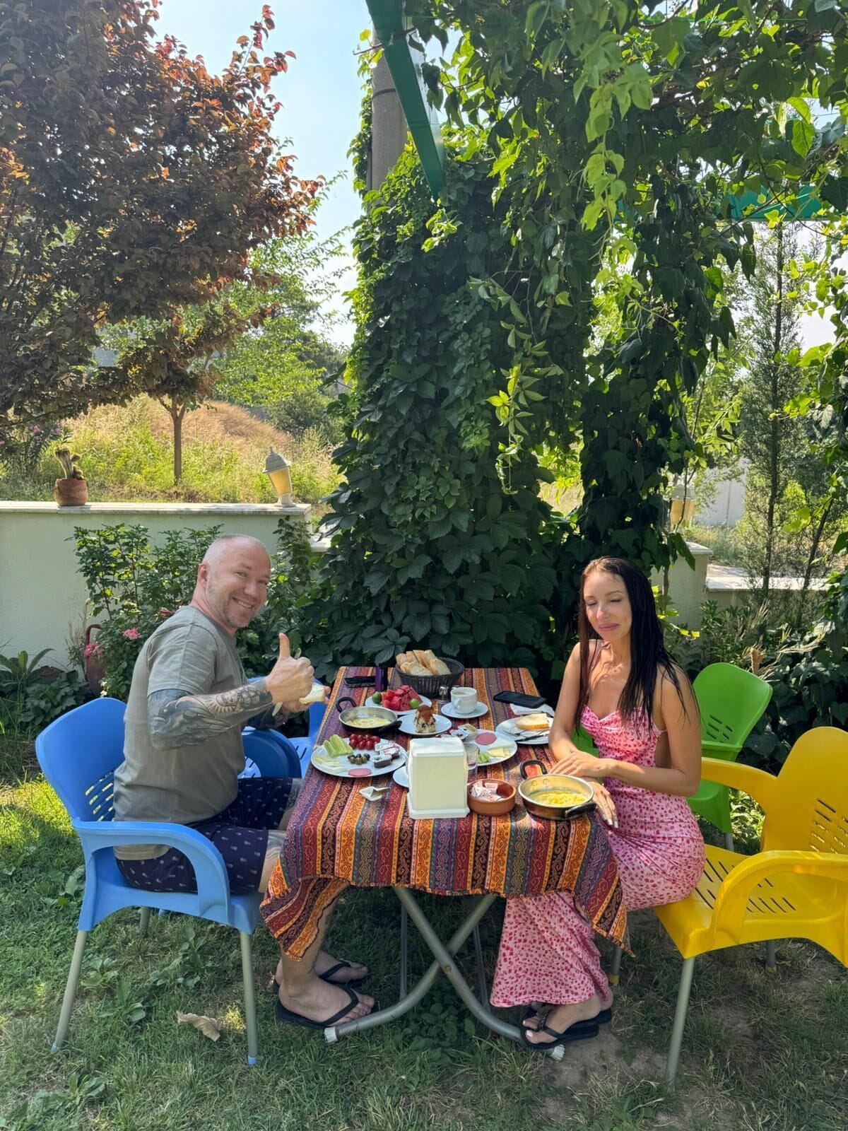 Two people seated at an outdoor table with various food items, surrounded by greenery and a clear sky.