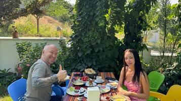 Two people seated at an outdoor table with various food items, surrounded by greenery and a clear sky.