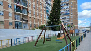 A playground with swings and slides, surrounded by a white wall and a tall building with balconies. - Sunny and central terrace apartment (Málaga)