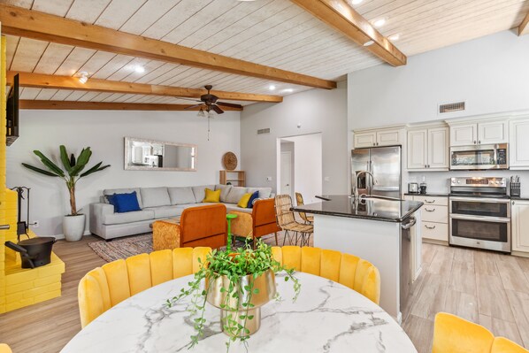 A modern kitchen with stainless steel appliances, a dining area with yellow chairs, and a living room with a gray sofa and a ceiling fan. - Scottsdale Game House Heated Pool, Golf, Bowling (Scottsdale)