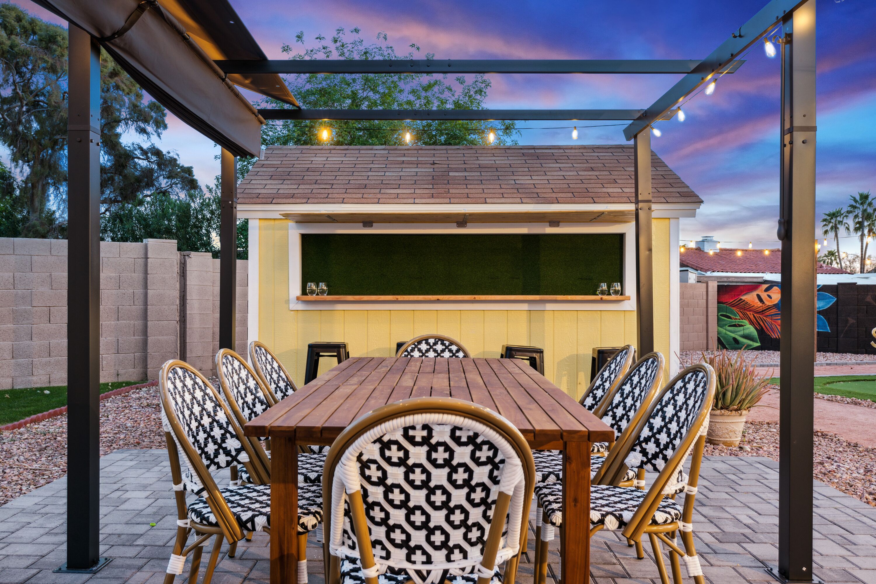 An outdoor patio with a wooden table and chairs, a pergola with string lights, and a mural on the wall.