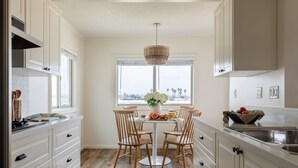 A kitchen with white cabinetry, a dining table with chairs, a chandelier, and a window with a view of palm trees.
