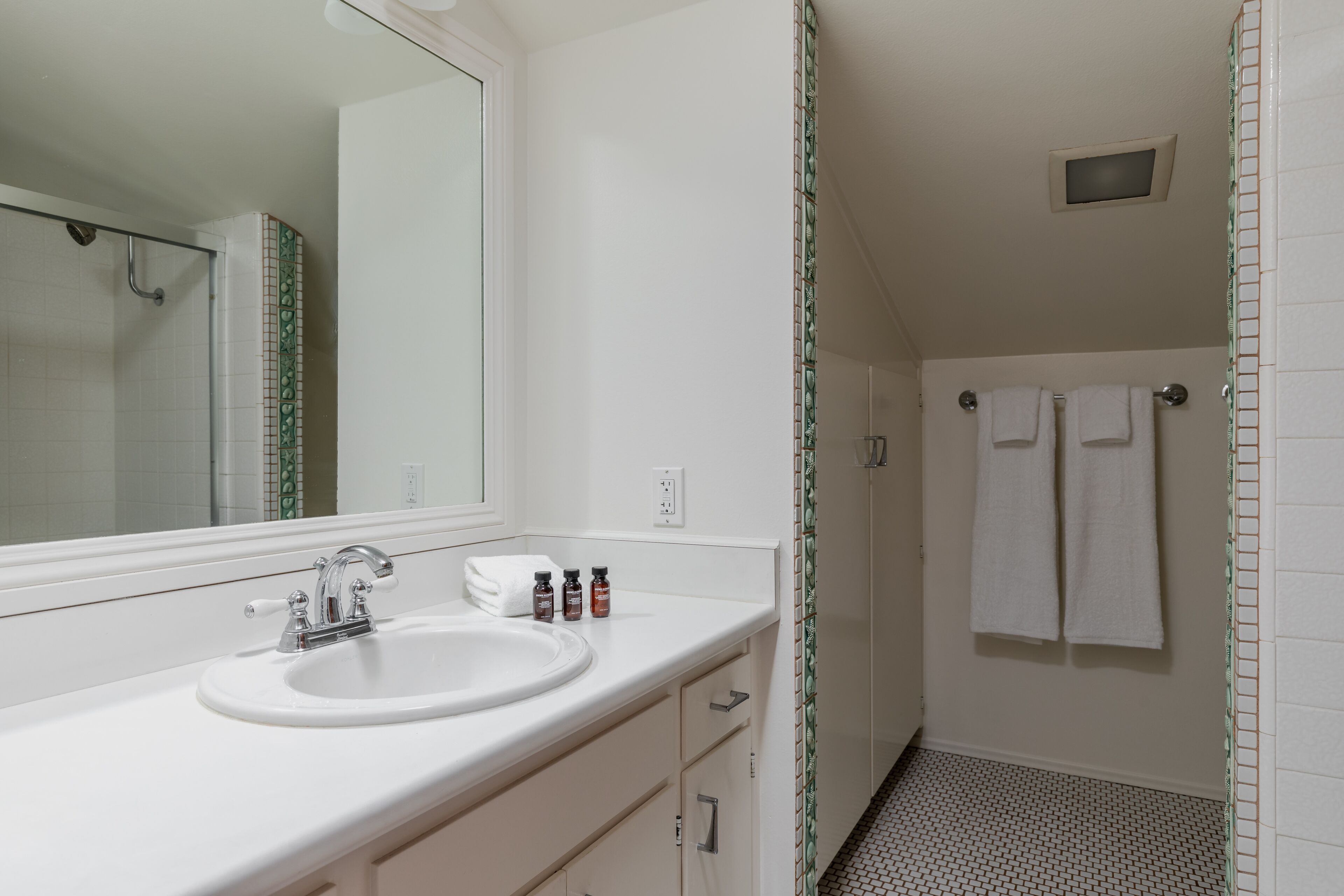 Une salle de bain avec un lavabo blanc, un miroir et un sol carrelé. Deux serviettes sont suspendues à un porte-serviettes.