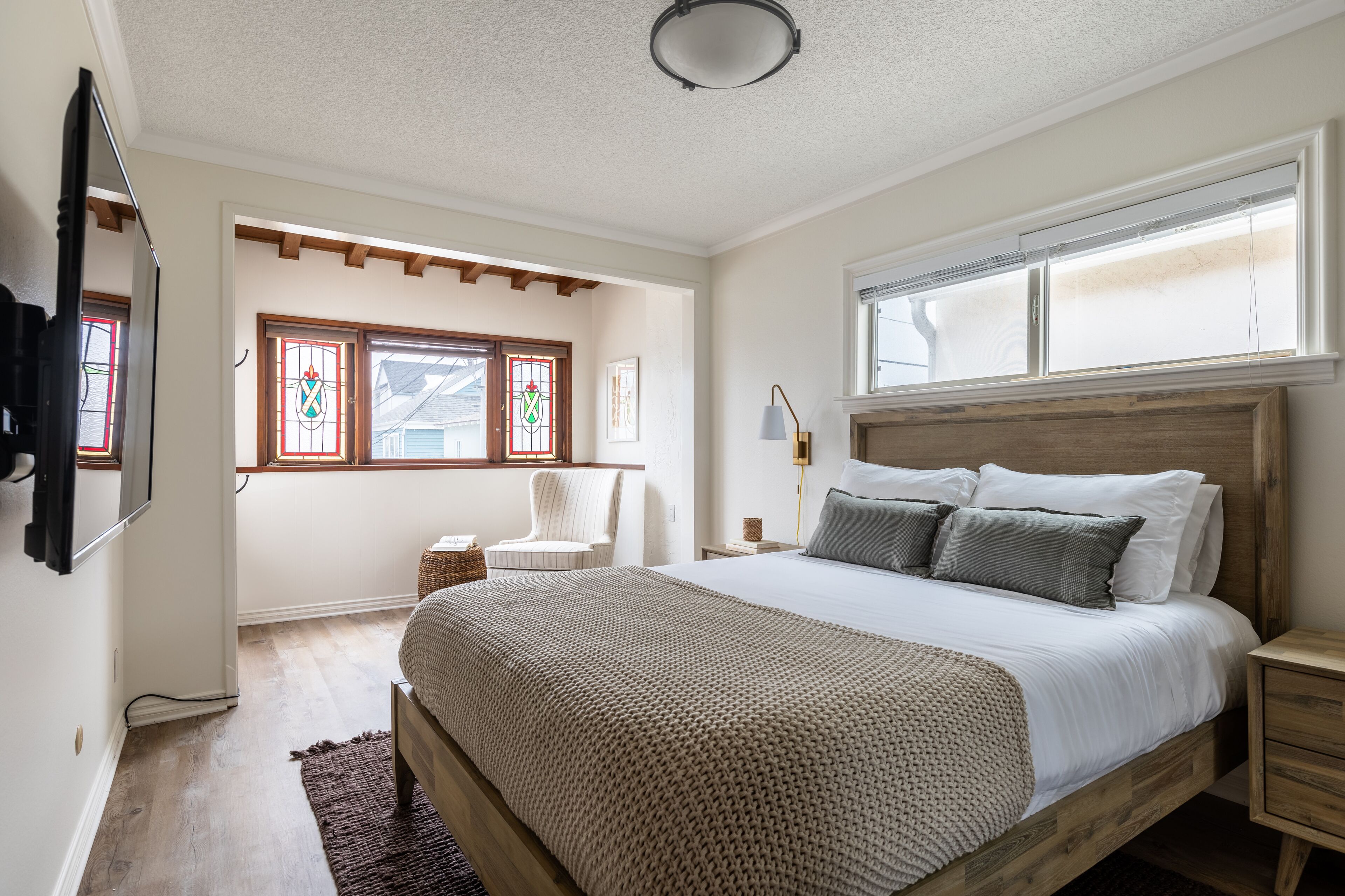 A bedroom with a wooden bed, a chair, a bedside table, and a window with stained glass.