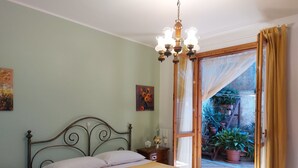 Une chambre à coucher avec un lit, une table de chevet, un lustre et une vue sur un jardin, visible à travers une porte ouverte.