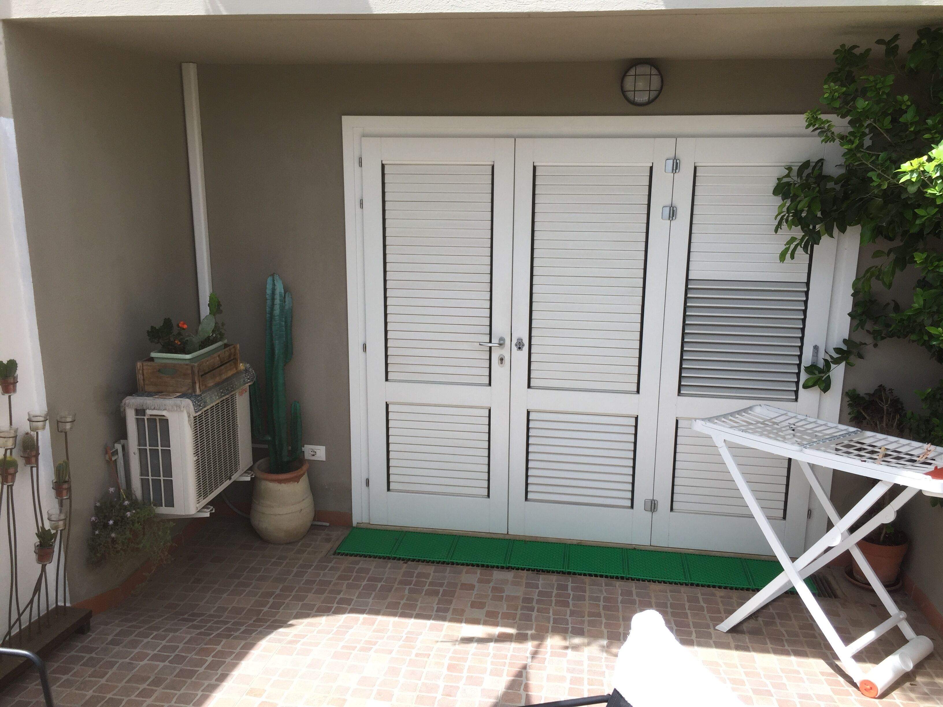 A patio with a white table and chairs, a green mat, a white door with shutters, and potted plants.