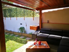 Un área de estar al aire libre con mesa de madera, cojines y una pérgola.