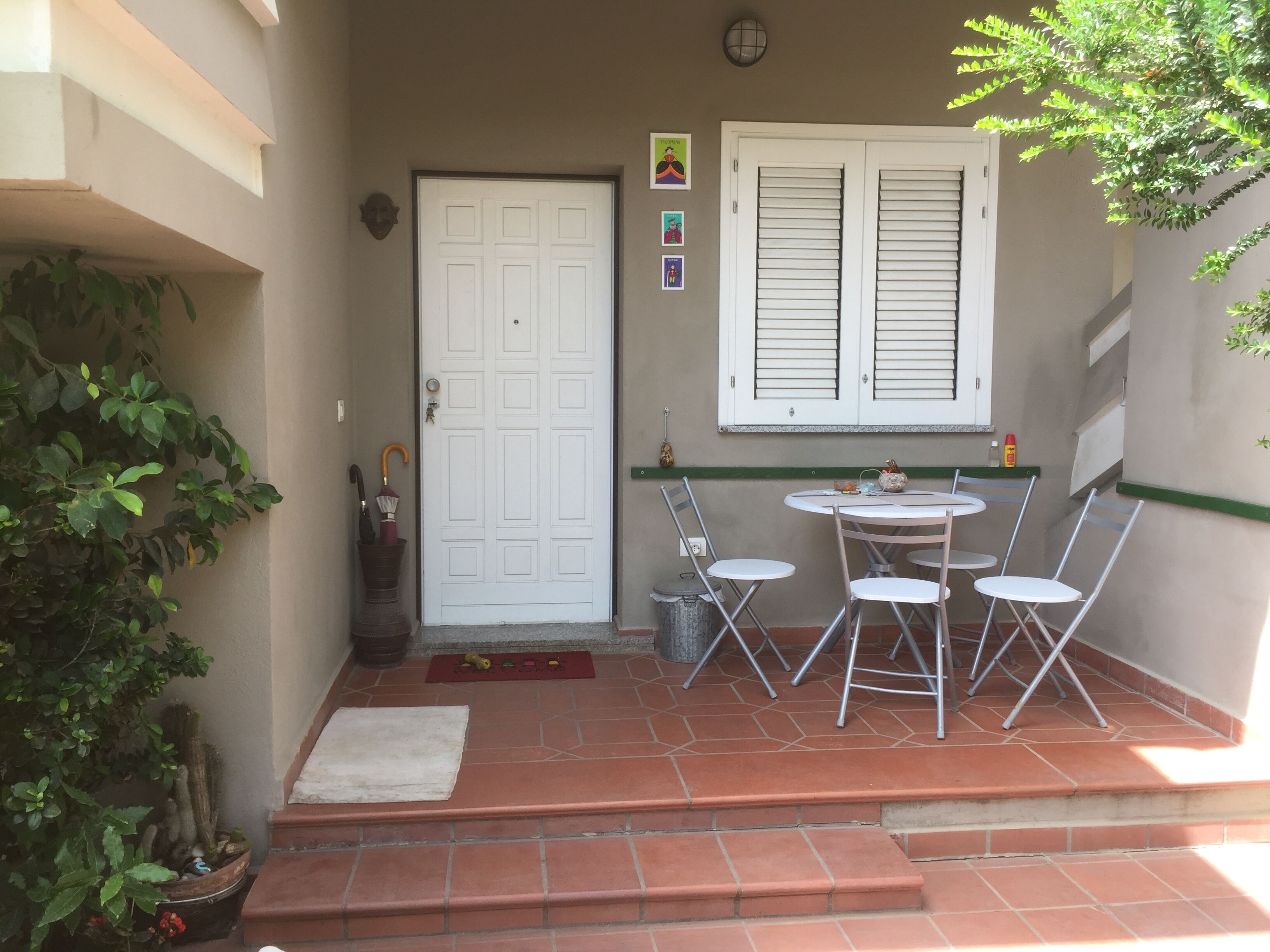 Un patio con una puerta blanca, una mesa pequeña con cuatro sillas y una ventana con contraventanas.