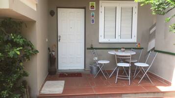 Un patio con una puerta blanca, una mesa pequeña con cuatro sillas y una ventana con contraventanas.