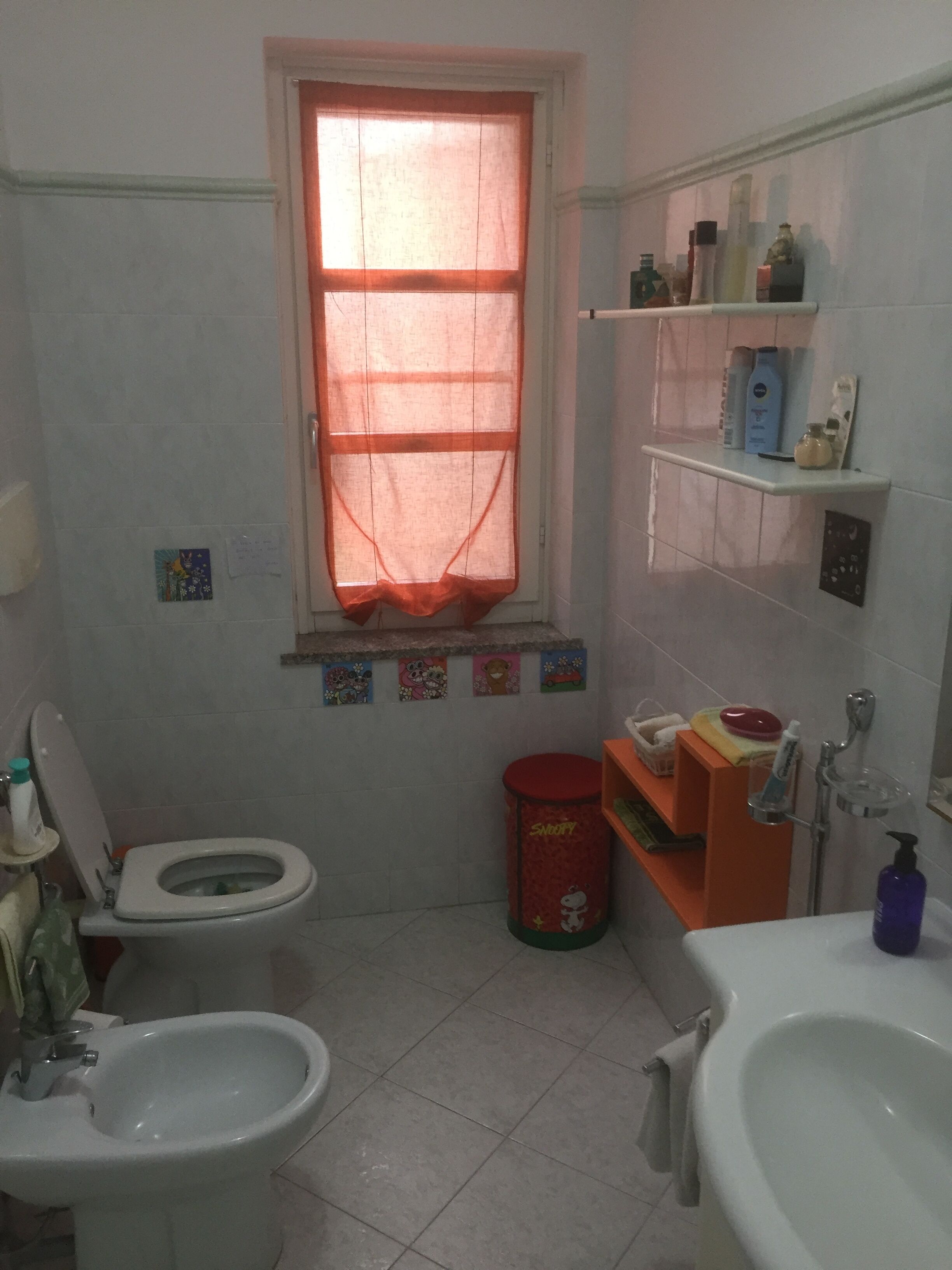 Ein kleines Badezimmer mit Toilette, Bidet und Waschbecken. Es gibt ein Fenster mit rotem Vorhang, ein Ablagefach mit Toilettenartikeln und einen Mülleimer.