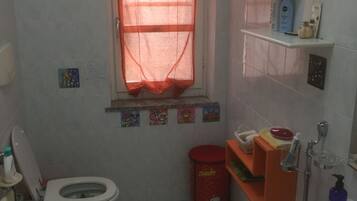 Ein kleines Badezimmer mit Toilette, Bidet und Waschbecken. Es gibt ein Fenster mit rotem Vorhang, ein Ablagefach mit Toilettenartikeln und einen Mülleimer.