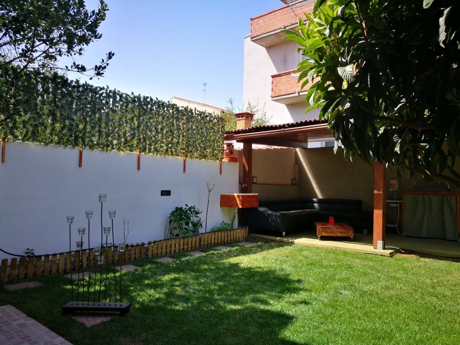 A covered outdoor seating area with a black sofa and a wooden table, surrounded by a white wall with a climbing plant.