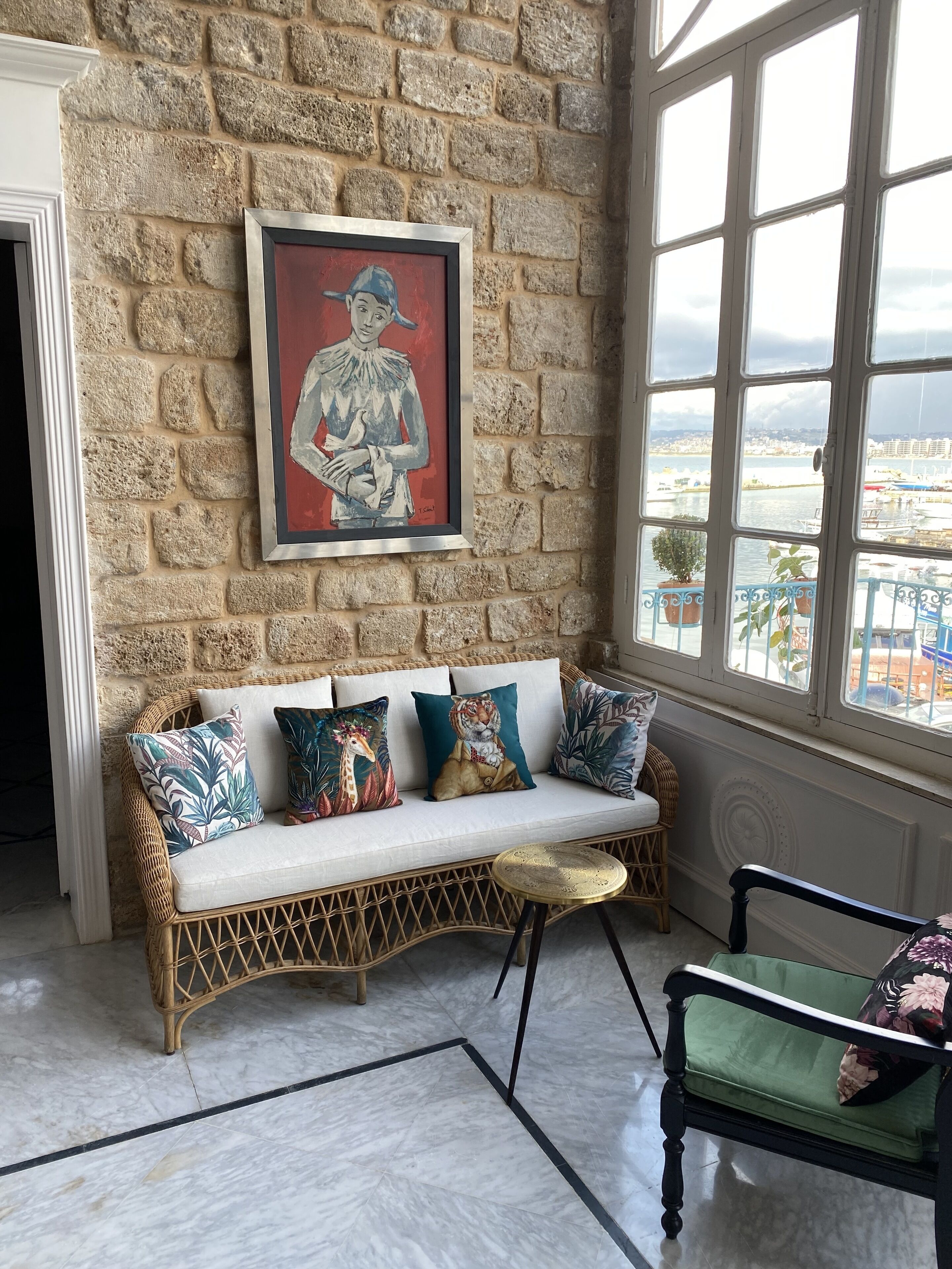 A wicker sofa with colorful cushions, a framed artwork on a stone wall, and a view of a harbor through large windows.