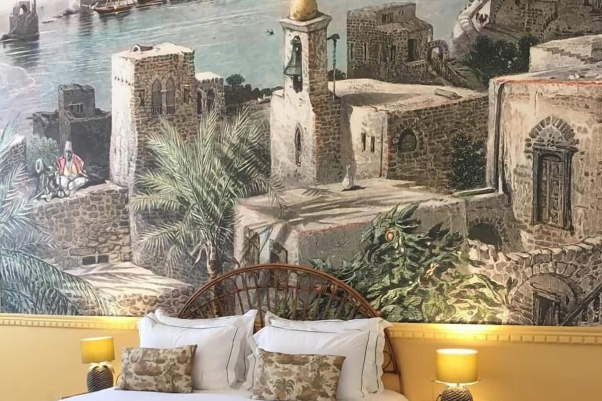 A bed with a view of a coastal town mural, a small bedside table with a lamp, and a decorative wall.