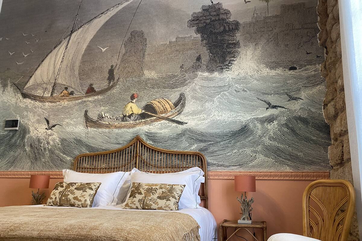 A bedroom with a large mural of a seascape, a bed with white and patterned pillows, a wicker chair, and a small table with a lamp.