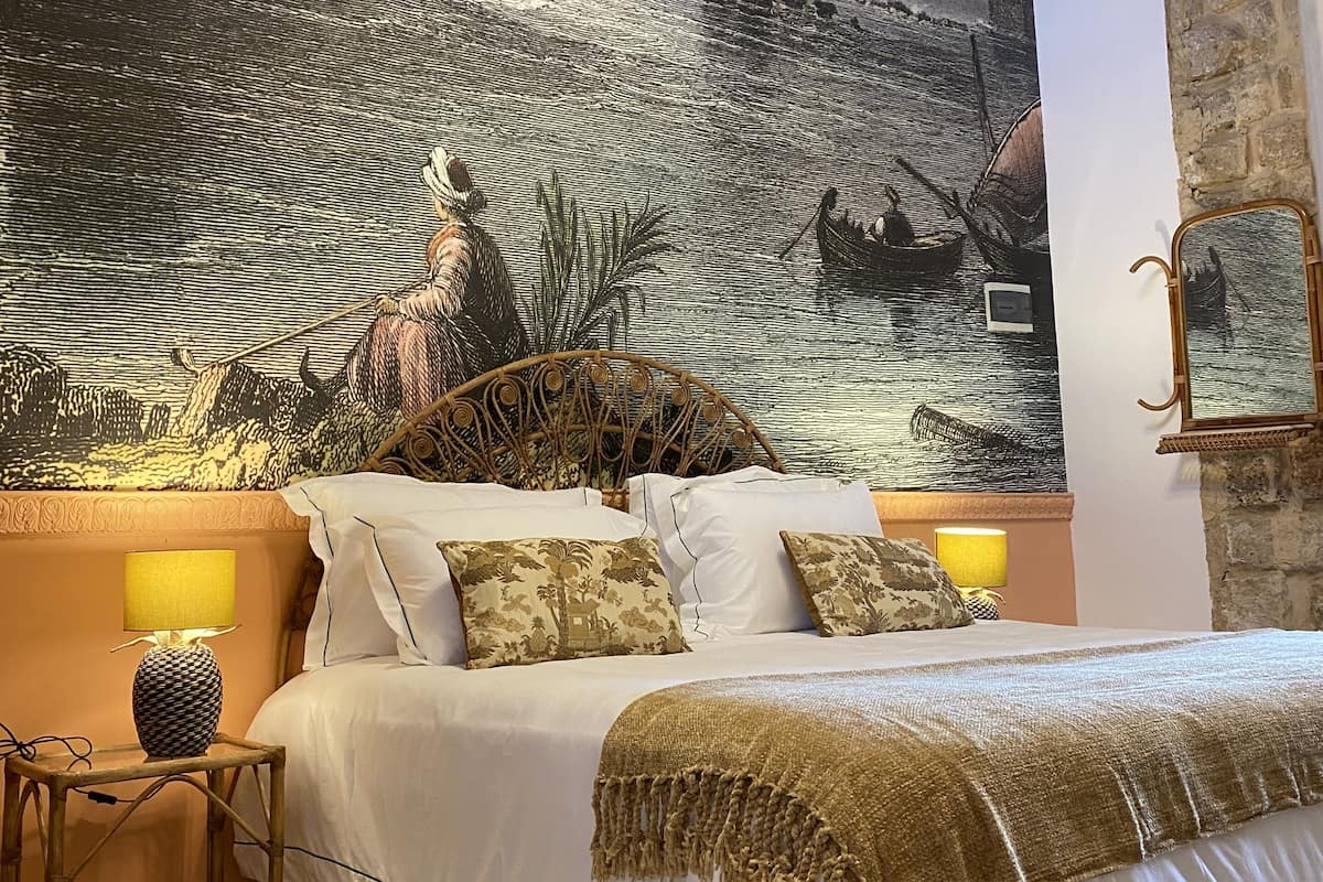 A bedroom with a large mural of a river scene, a bed with white linens, a wooden headboard, and two bedside lamps.