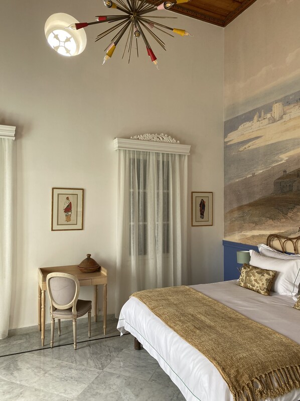 A bedroom with a bed, a desk, and a chair. There is a painting on the wall and a window with curtains. - Dar Alice (Tyre)