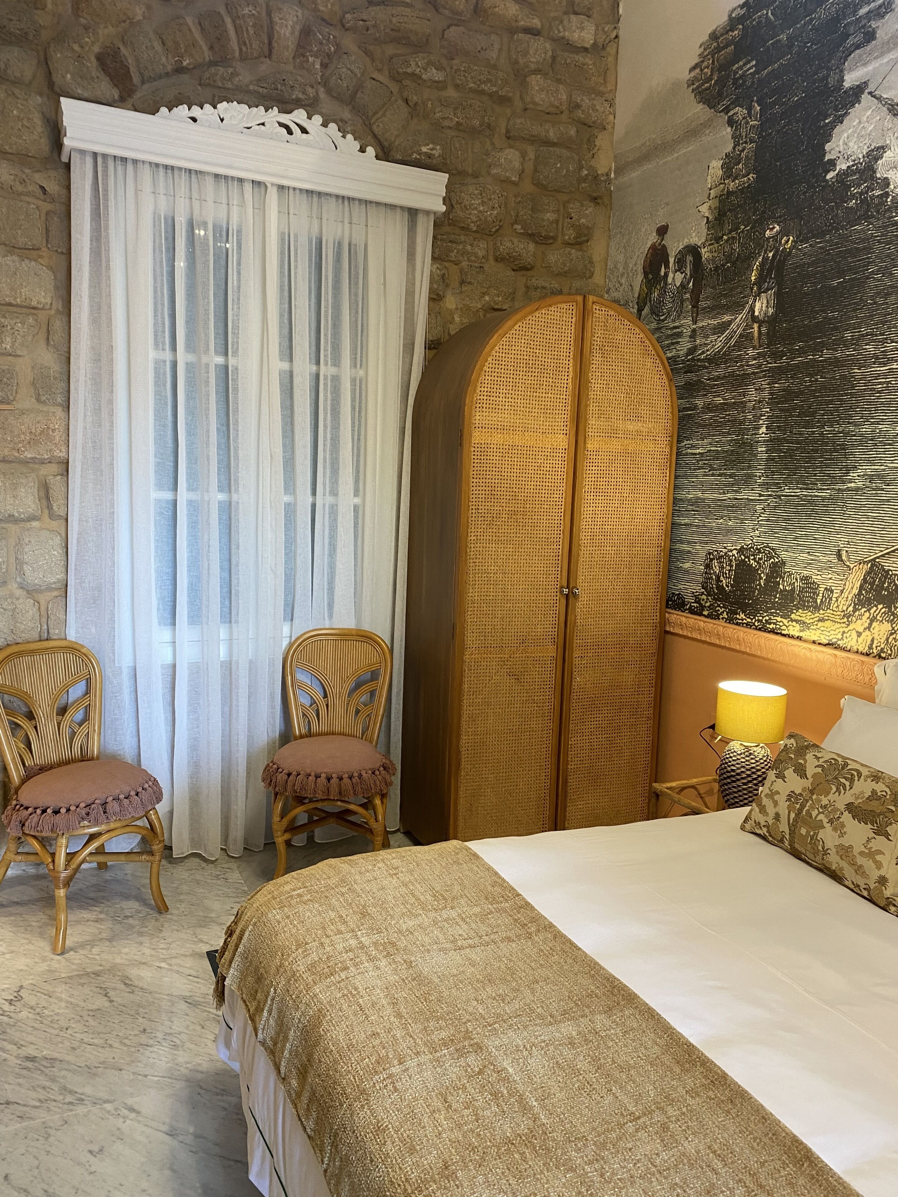 A room with a bed, two wicker chairs, a rattan wardrobe, a large tapestry, and a stone wall.