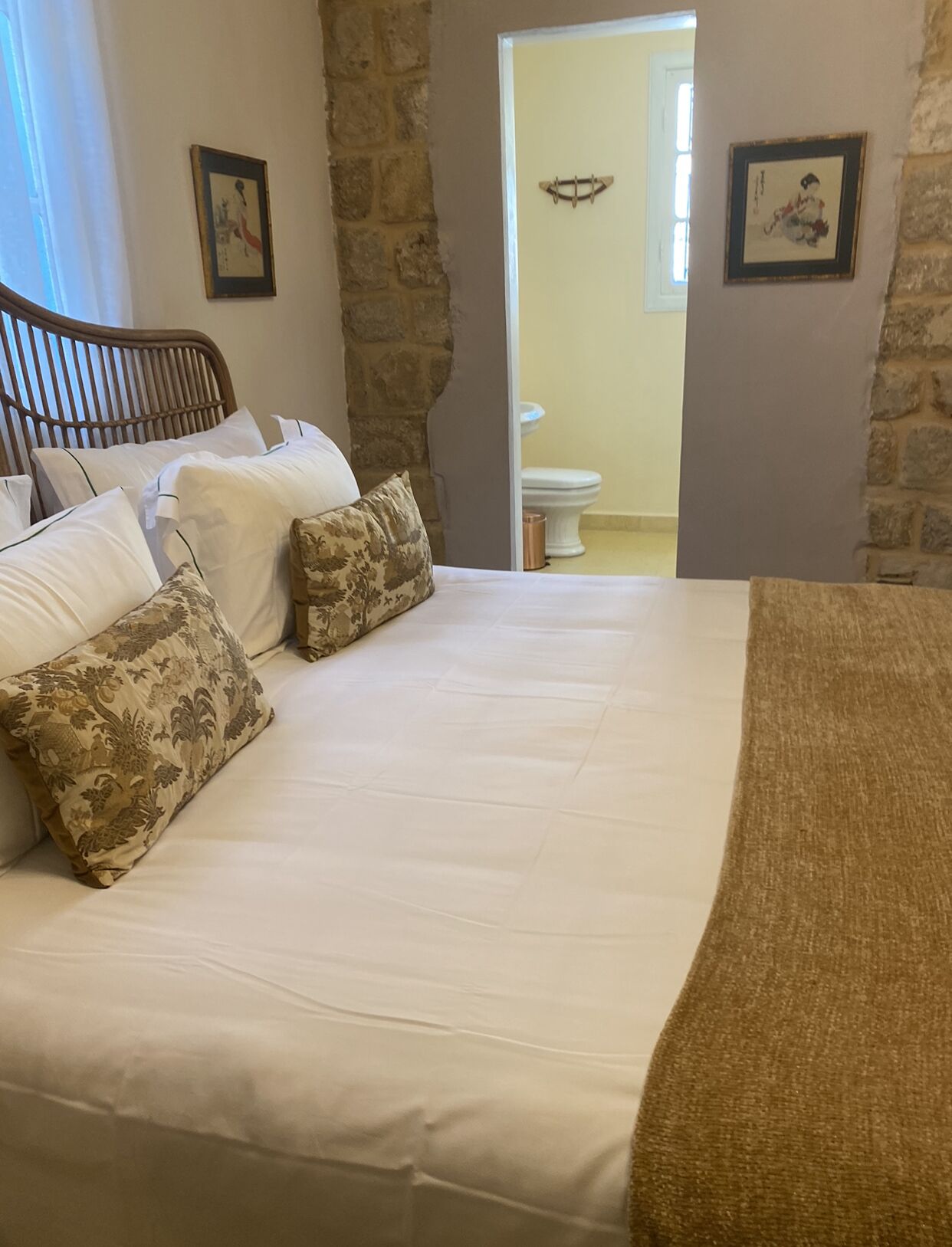 Una cama bien hecha con sábanas blancas y almohadas estampadas, cabecera de madera y pared de piedra.