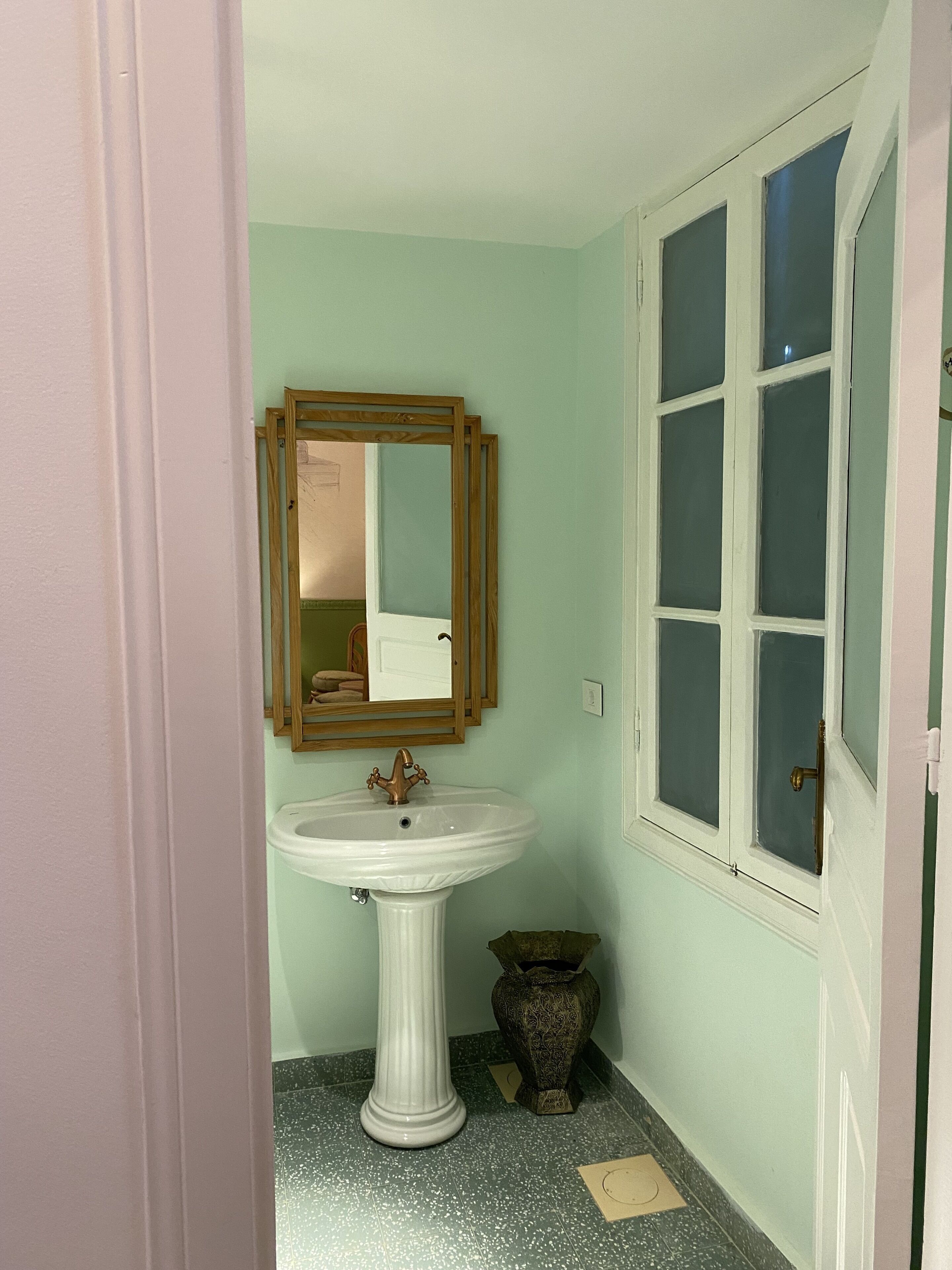 Un baño con lavamanos pedestal, espejo, ventana y puerta.
