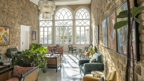 A spacious room with a high ceiling, stone walls, and large arched windows allowing natural light. There are several seating areas with wicker furniture and a variety of plants. - Dar Alice (Tyre)