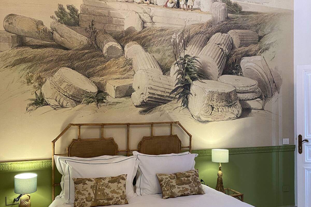 A bedroom with a large mural of ruins, a bed with patterned pillows, and two bedside lamps.