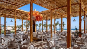 Een buitenruimte met een eethoek, houten pergola’s, witte rieten meubels en uitzicht op het strand.