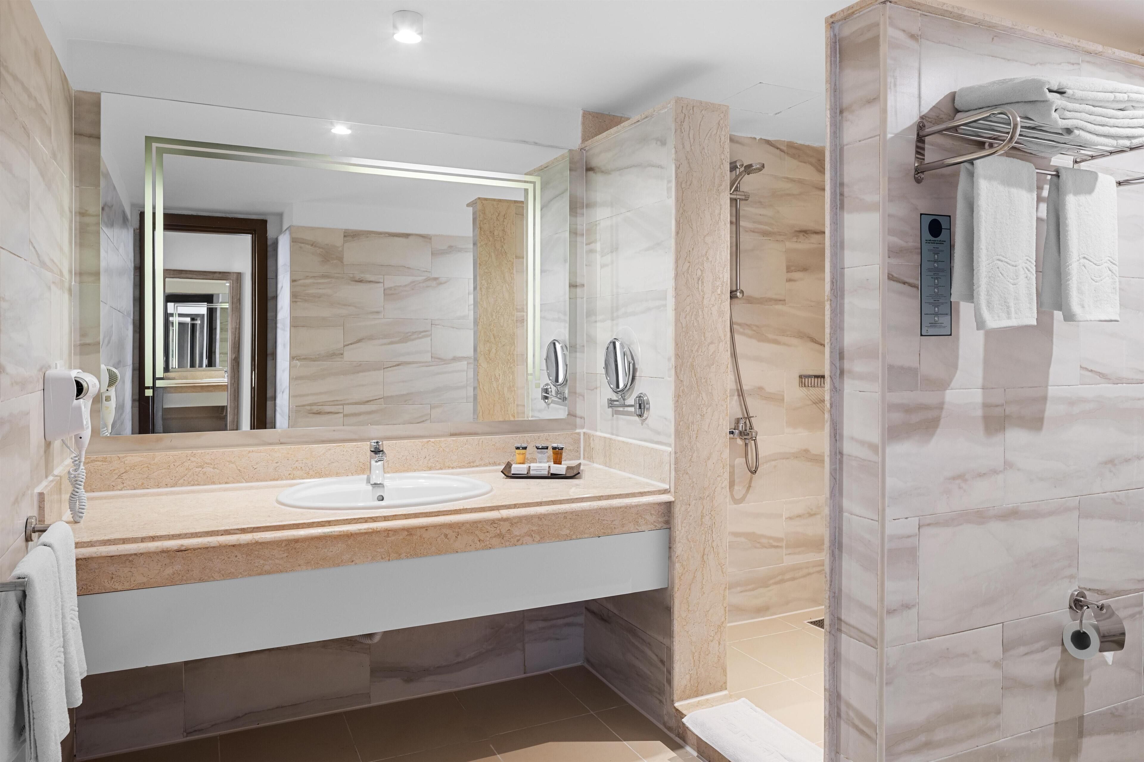 A modern bathroom with a marble vanity, a large mirror, a shower with a handheld showerhead, and a towel rack with folded towels.