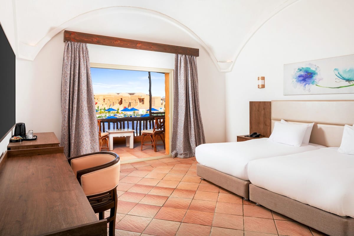 Een hotelkamer met een bed, een bureau en een stoel. Er is uitzicht op een zwembad en gebouwen buiten het raam.