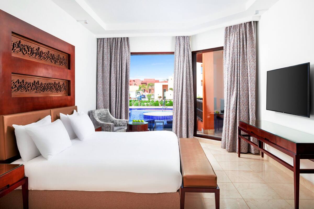 Hotelkamer met een groot bed, televisie, bureau en uitzicht op een zwembad en gebouwen.