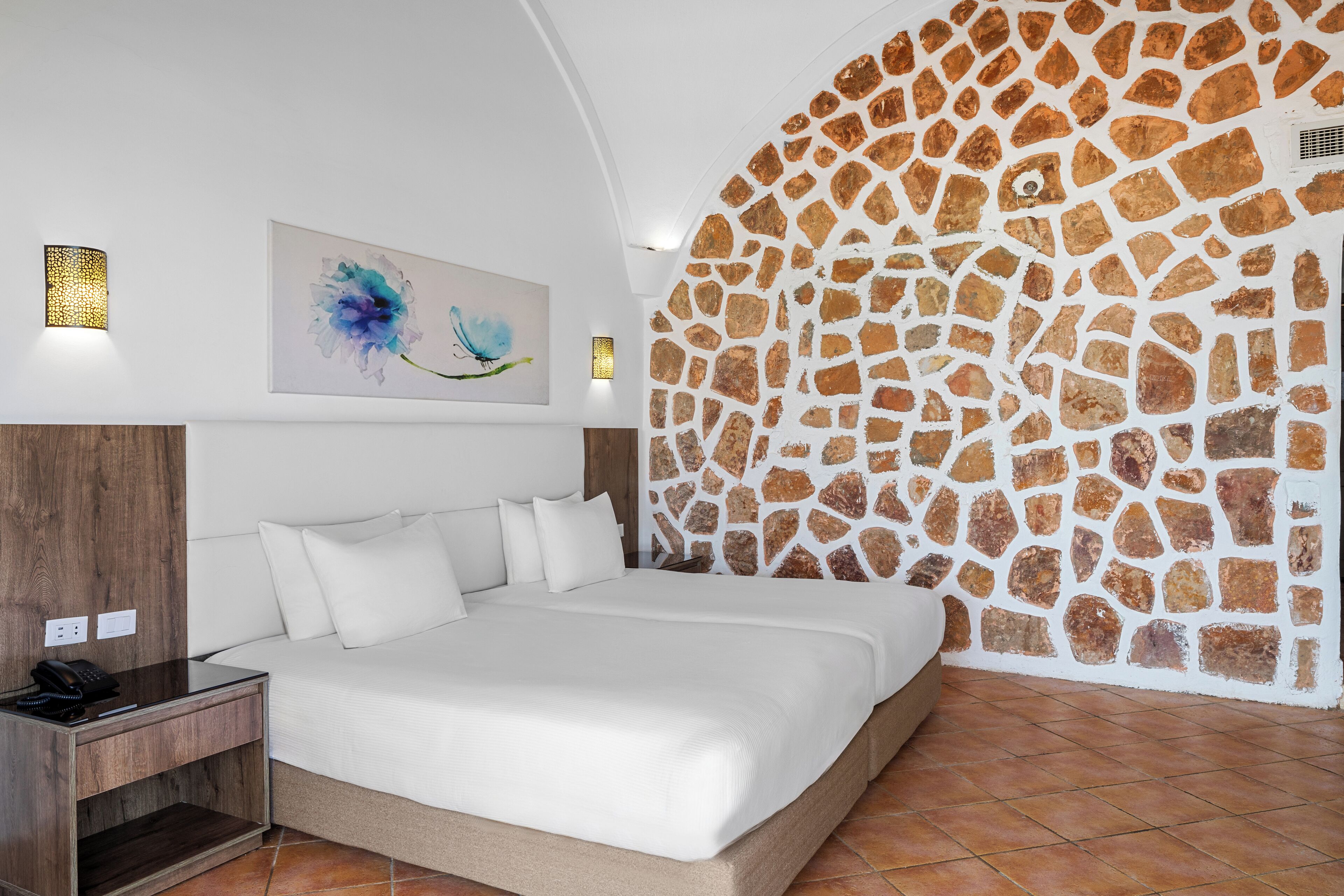 A modern bedroom with a stone wall, a bed with white linens, a nightstand, and a wall-mounted lamp.