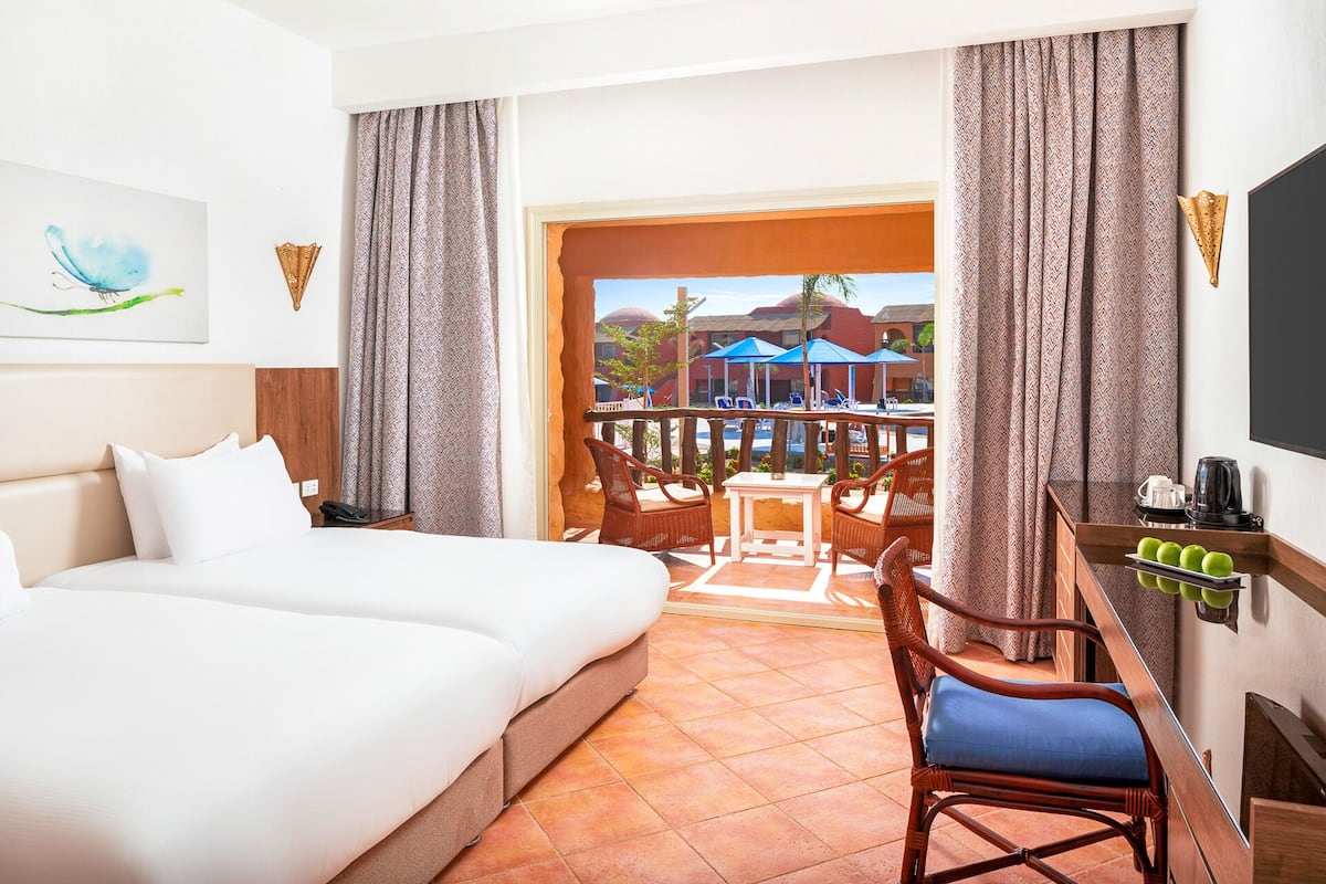 Een hotelkamer met een bed, een bureau met stoel, een televisie en een balkon met zwembad en parasols.