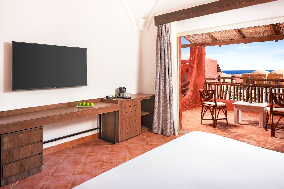 Een kamer met een bed, een bureau met een flatscreen-televisie, een koffiezetapparaat en uitzicht op zee vanaf een balkon met rieten meubilair.
