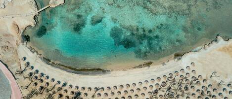 An aerial view of a beach with clear turquoise waters, a sandy shoreline, and numerous umbrellas arranged in rows.