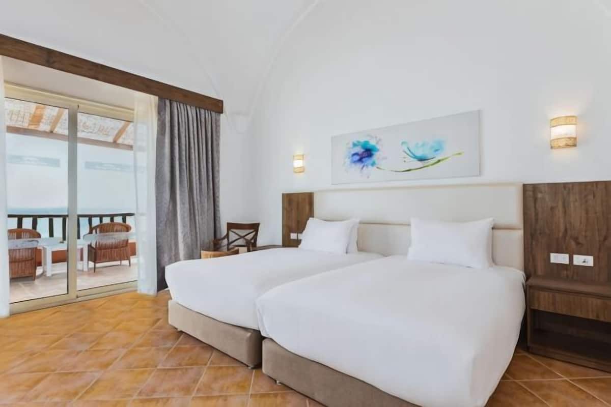 Een hotelkamer met twee bedden, een balkon met rieten meubilair en een schilderij aan de muur.