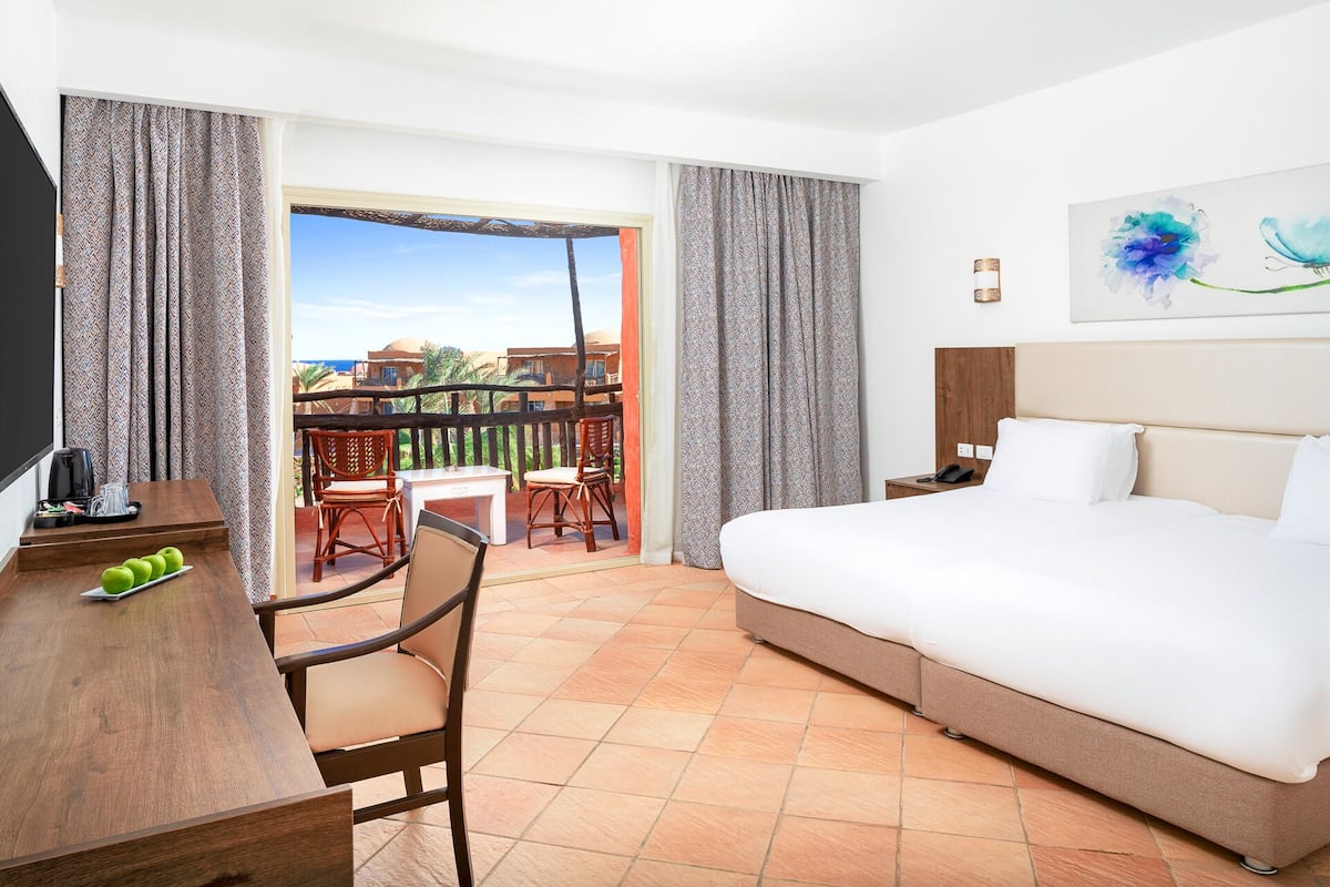 Een hotelkamer met een bed, een bureau met stoelen, een balkon met uitzicht op gebouwen en palmbomen, en een schilderij aan de muur.