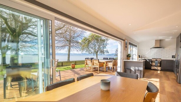 Una cocina moderna con una isla grande, una zona de comedor con una mesa de madera y vistas a un lago a través de puertas de vidrio amplias.