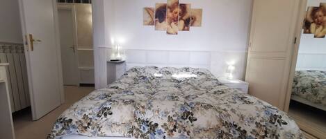 Um quarto com colcha florida, cabeceira branca, mesa de cabeceira com abajur e espelho na parede.
