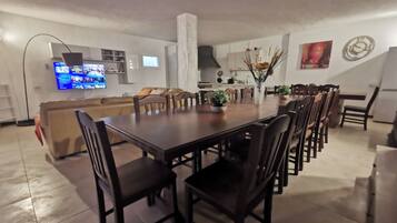 Un comedor amplio con una mesa larga y numerosas sillas, un televisor montado en la pared y una cocina al fondo.