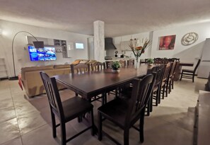 Un comedor amplio con una mesa larga y numerosas sillas, un televisor montado en la pared y una cocina al fondo.