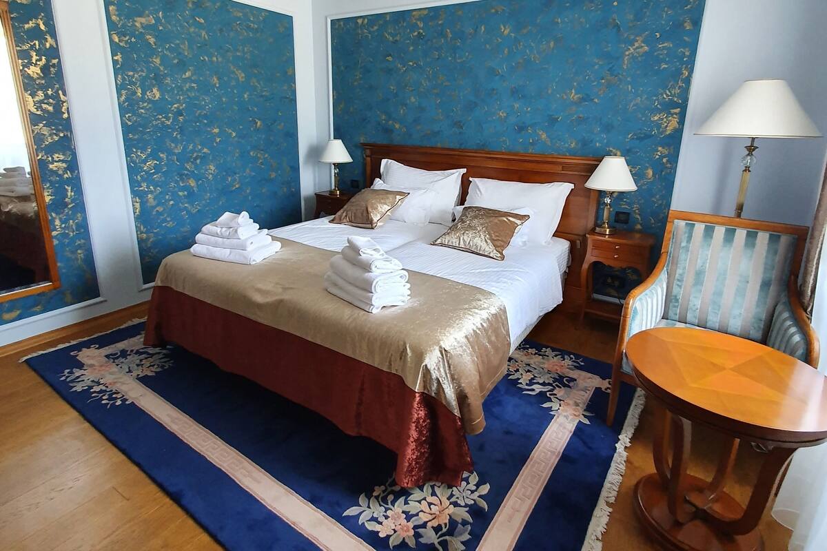 Ein Schlafzimmer mit einem Bett, zwei Nachttischen, einem Sessel, einem großen Teppich und blauen Wänden mit goldenen Akzenten.