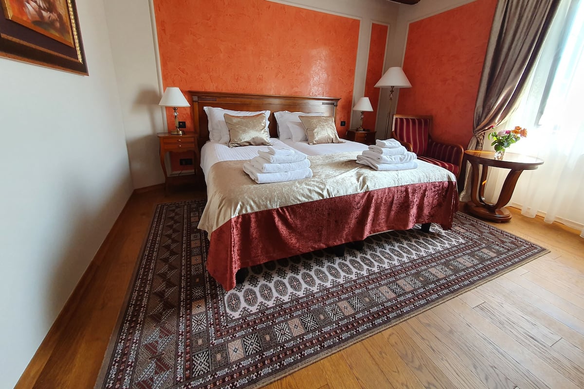 Ein ordentlich bezogenes Bett mit weißen Leinentüchern und einer roten Tagesdecke, ein Nachttisch aus Holz mit einer Lampe und ein gemustertes Teppich auf Holzdielen.