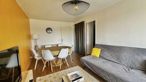 A compact living space with a sofa, dining table, chairs, a TV, and a patterned rug. - Capucine, Duplex à 5 min de la mer (Courseulles-sur-Mer)