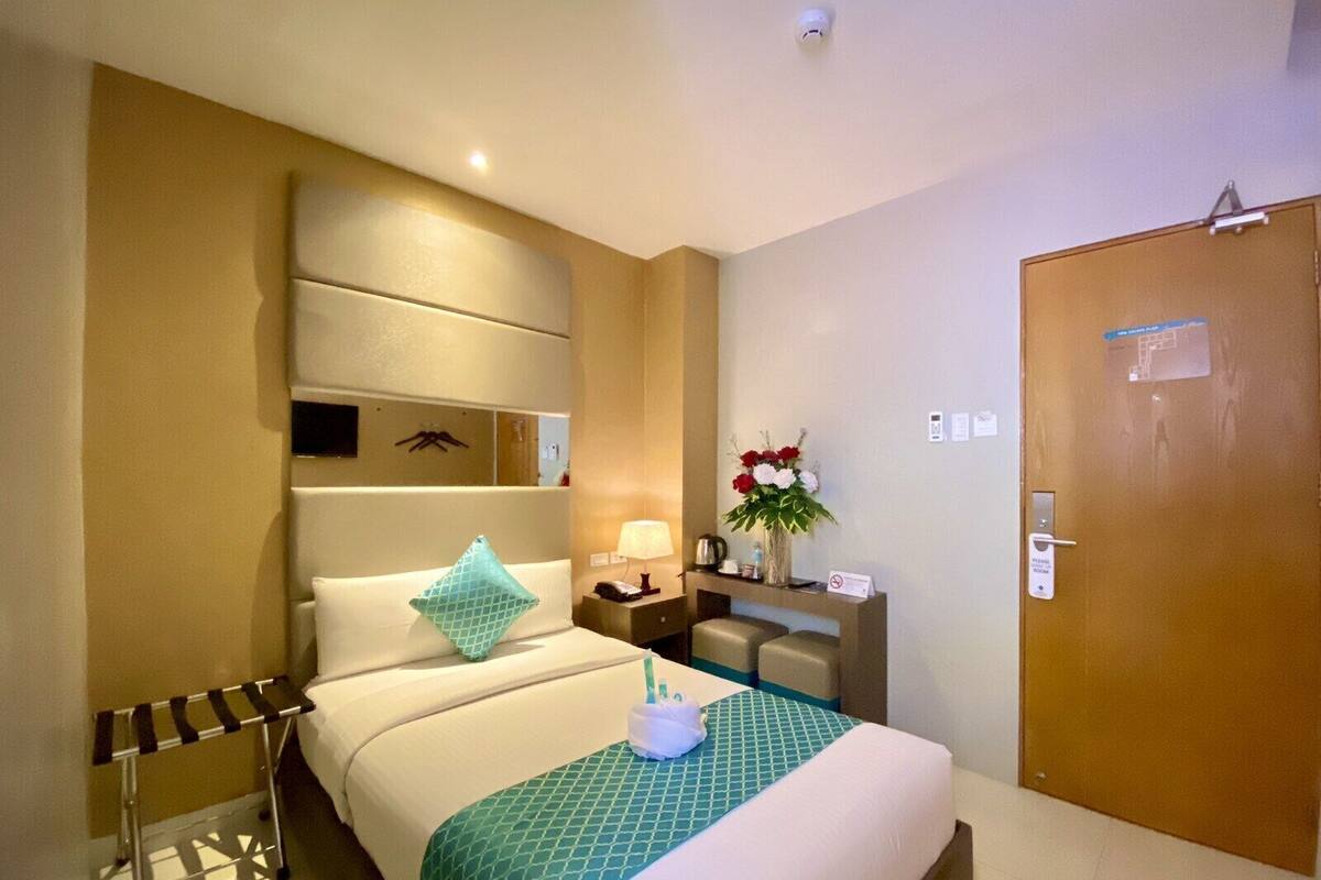 Ein Hotelzimmer mit einem Bett, einem Nachttisch mit Lampe, einem kleinen Schreibtisch, einem Sessel und einer Holztür.