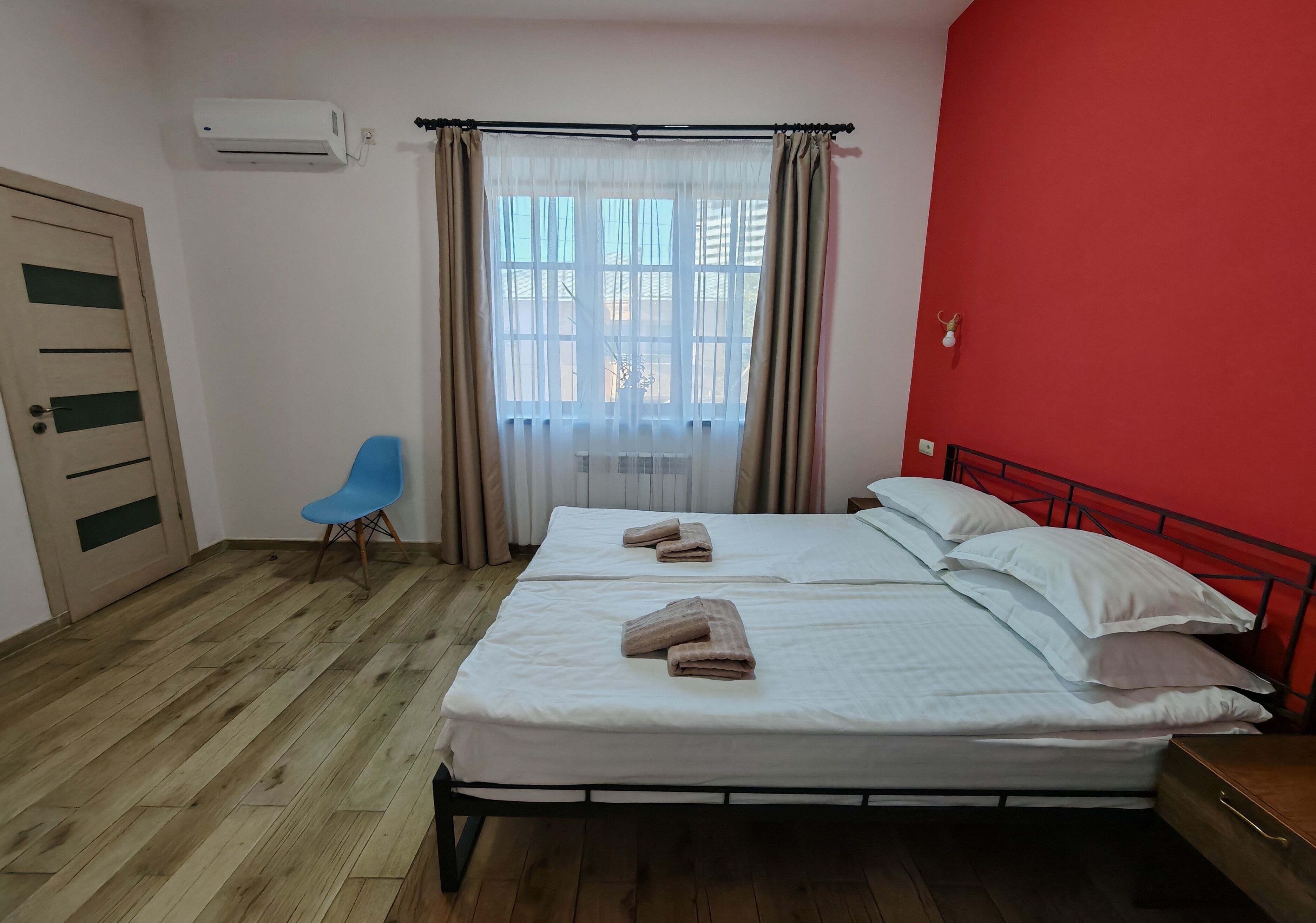 A bedroom with two beds, a blue chair, a wooden floor, a red wall, and a window with curtains.