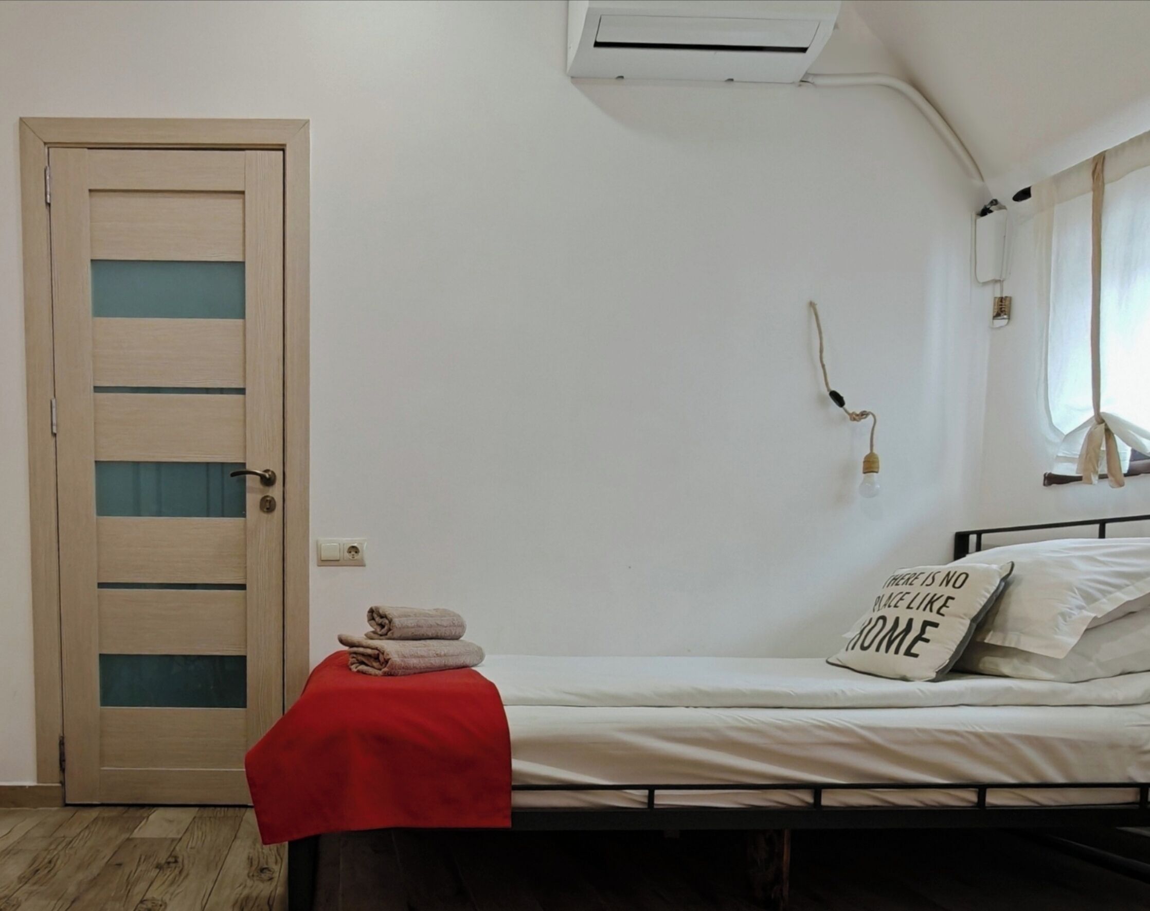 A small, single-bed room with a wooden door, a red towel on the floor, a wall-mounted air conditioner, and a bedside lamp.