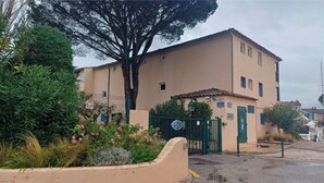 Un edificio beige con un ingresso sorvegliato da un cancello, immerso nel verde e con un albero di grandi dimensioni.