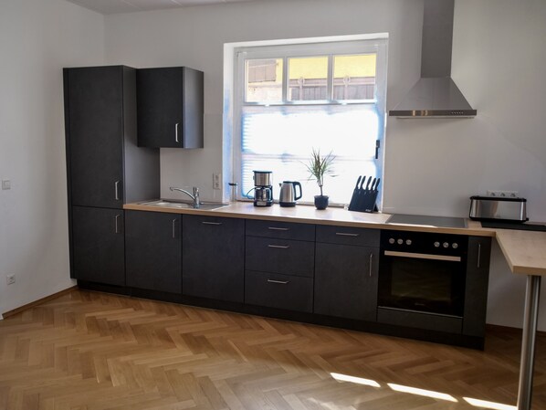 Une cuisine moderne avec des meubles de couleur sombre, un plan de travail en bois, un four encastré et une fenêtre donnant sur une clôture.