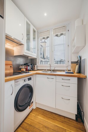 A compact kitchen with a washing machine, wooden countertop, white cabinets, and a window with a view.
